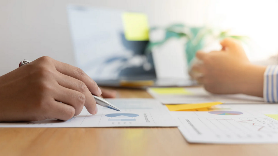 Image of two people consulting. You can only see their hands, and they are working on paperwork.